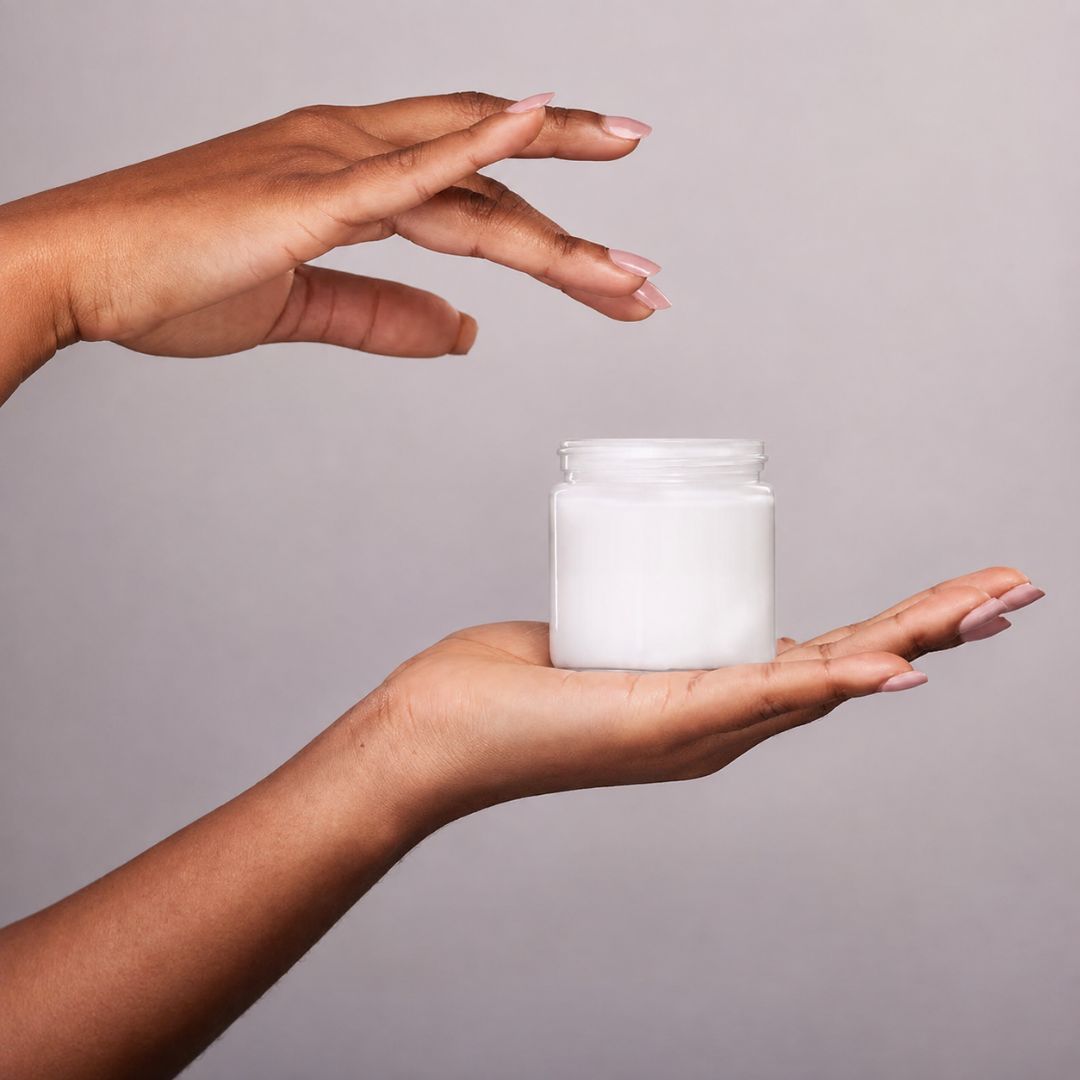 A person's hands holding an open white jar of skincare cream against a solid gray background.