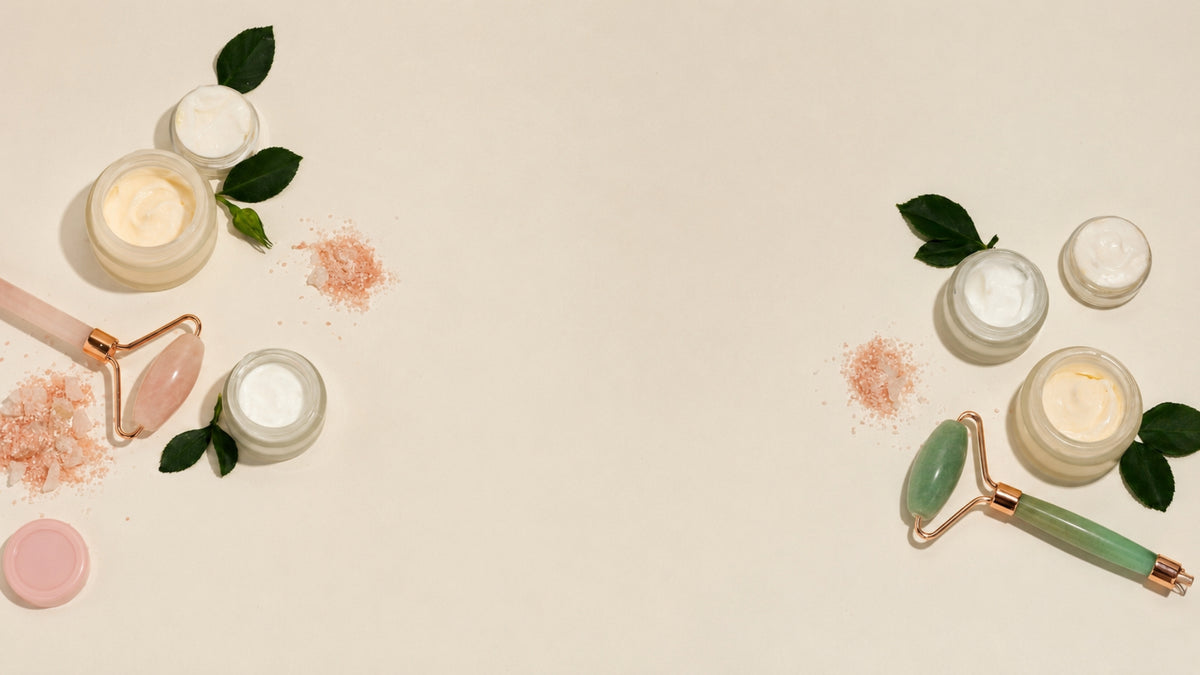 Two jade roller massage devices and several small jars of skincare cream are arranged on a light beige background with scattered rose powder and green leaves. The left set features a pink handle and rose gold metal, while the right set features a green handle and rose gold metal.