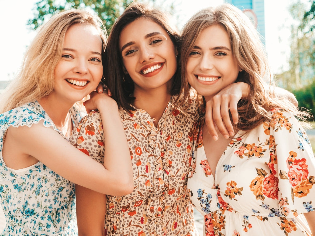 Three smiling women hugging outdoors, each wearing a different floral and patterned dress in blue, beige, and white with orange floral details.
