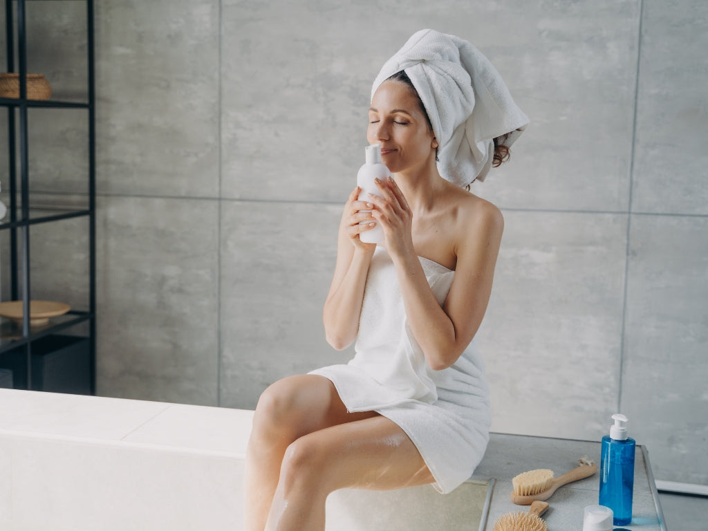 A woman wrapped in a white towel with a white towel turban on her head sits on a white bathtub in a modern bathroom, holding a white bottle. A blue bottle and wooden brushes are on the counter next to her.